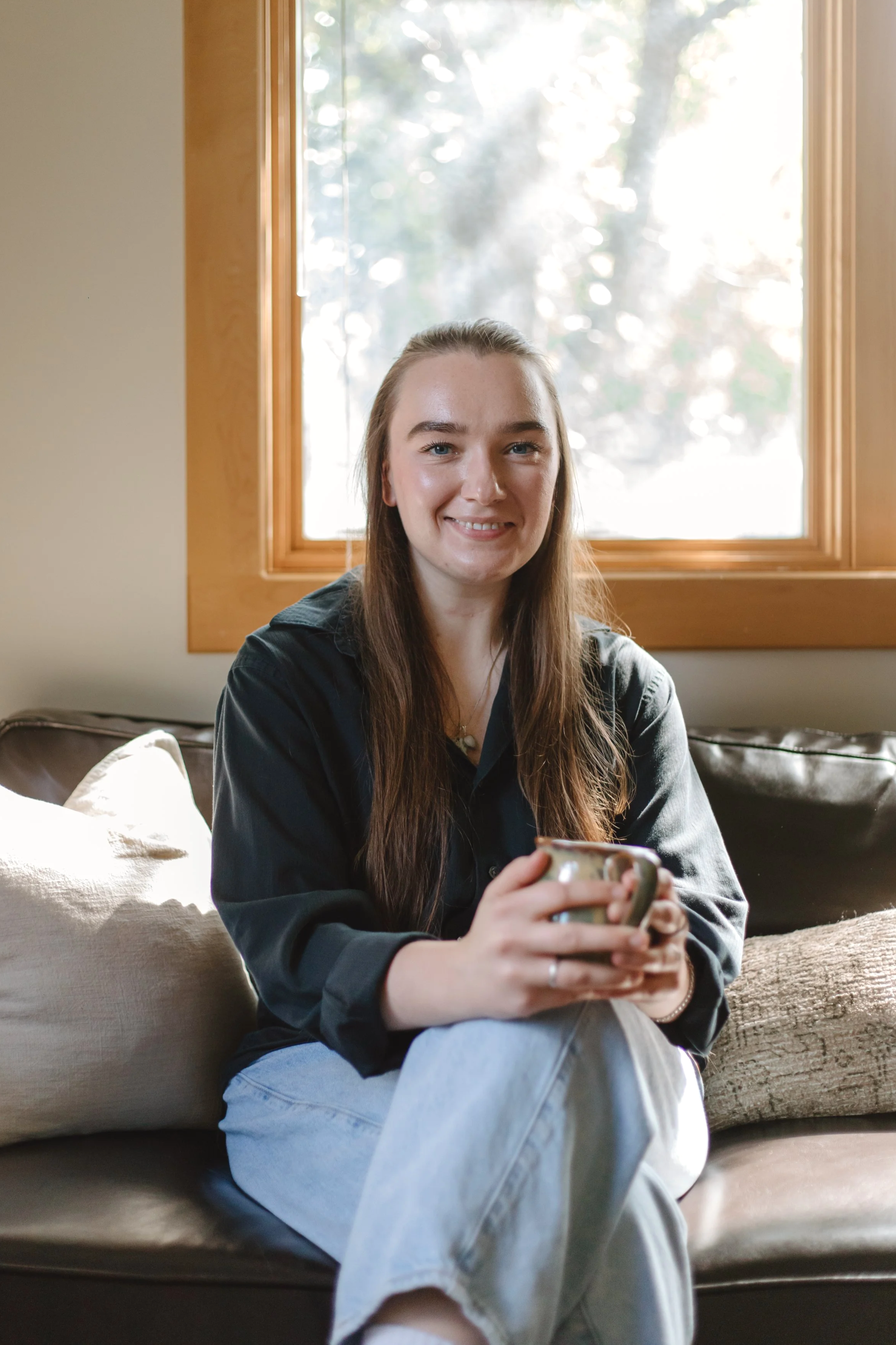 A young woman with long brown hair sitting on a beige couch by a window, holding a mug, smiling, with natural light coming through the window.