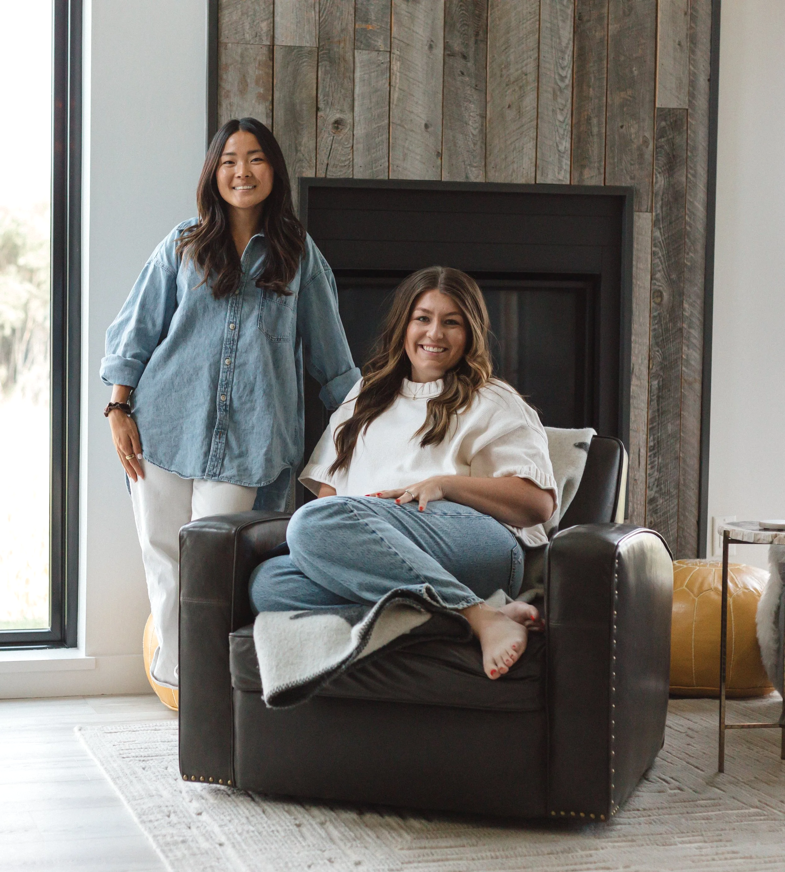 Two women in a living room with a modern dark gray armchair; one sitting with legs crossed and feet on the armrest, the other standing beside her. Both smiling, casually dressed, with a wooden accent wall behind them, large window to the side, and a yellow ottoman and side table nearby.