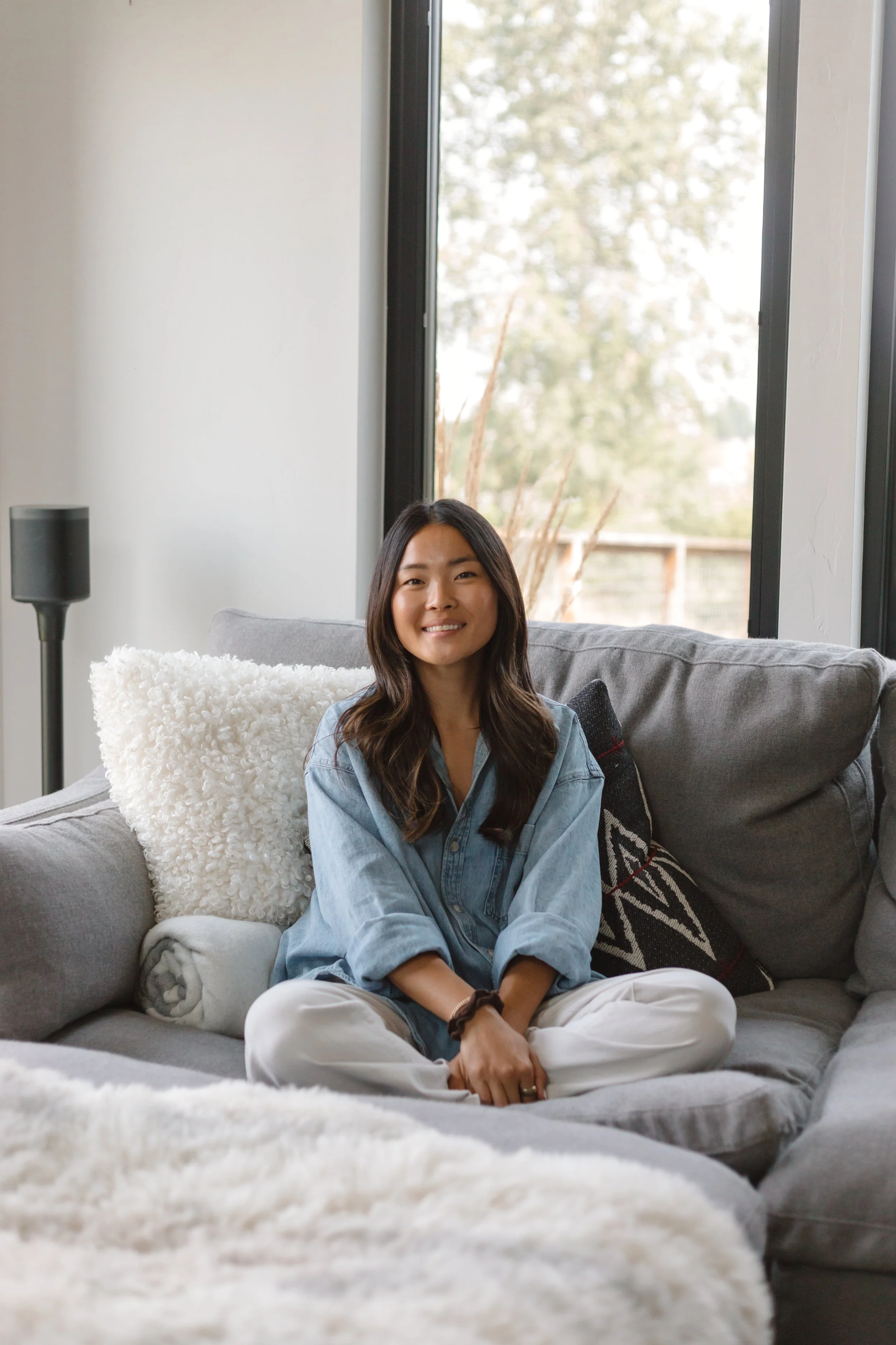 A woman sitting cross-legged on a gray sofa with a white fluffy pillow behind her, smiling at the camera. She wears a denim shirt and white pants, with long dark hair and a wristwatch. Behind her, a large window shows trees outside, and a black speaker is visible to the left.