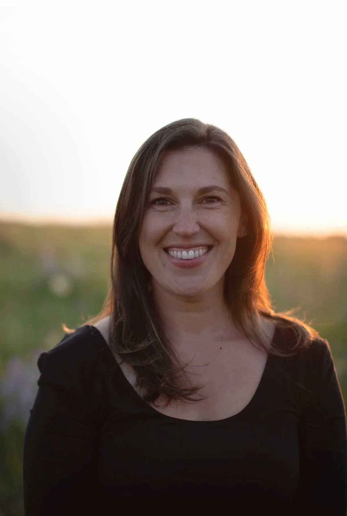 A woman with long brown hair smiling outdoors during sunset, wearing a black top.