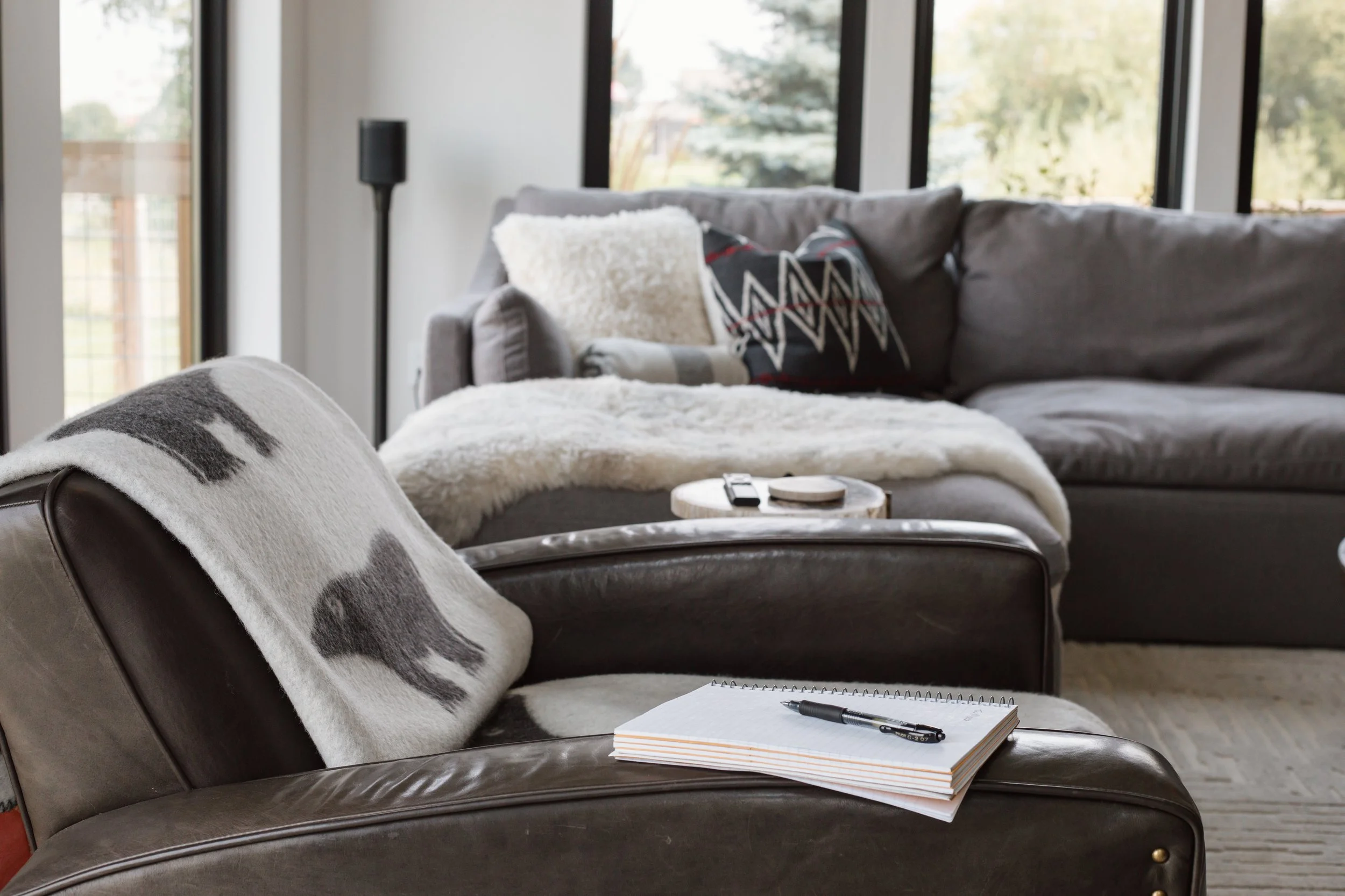 Living room with gray sofa, throw pillows, black side lamp, large windows with trees outside, notebook and pen on dark leather chair.