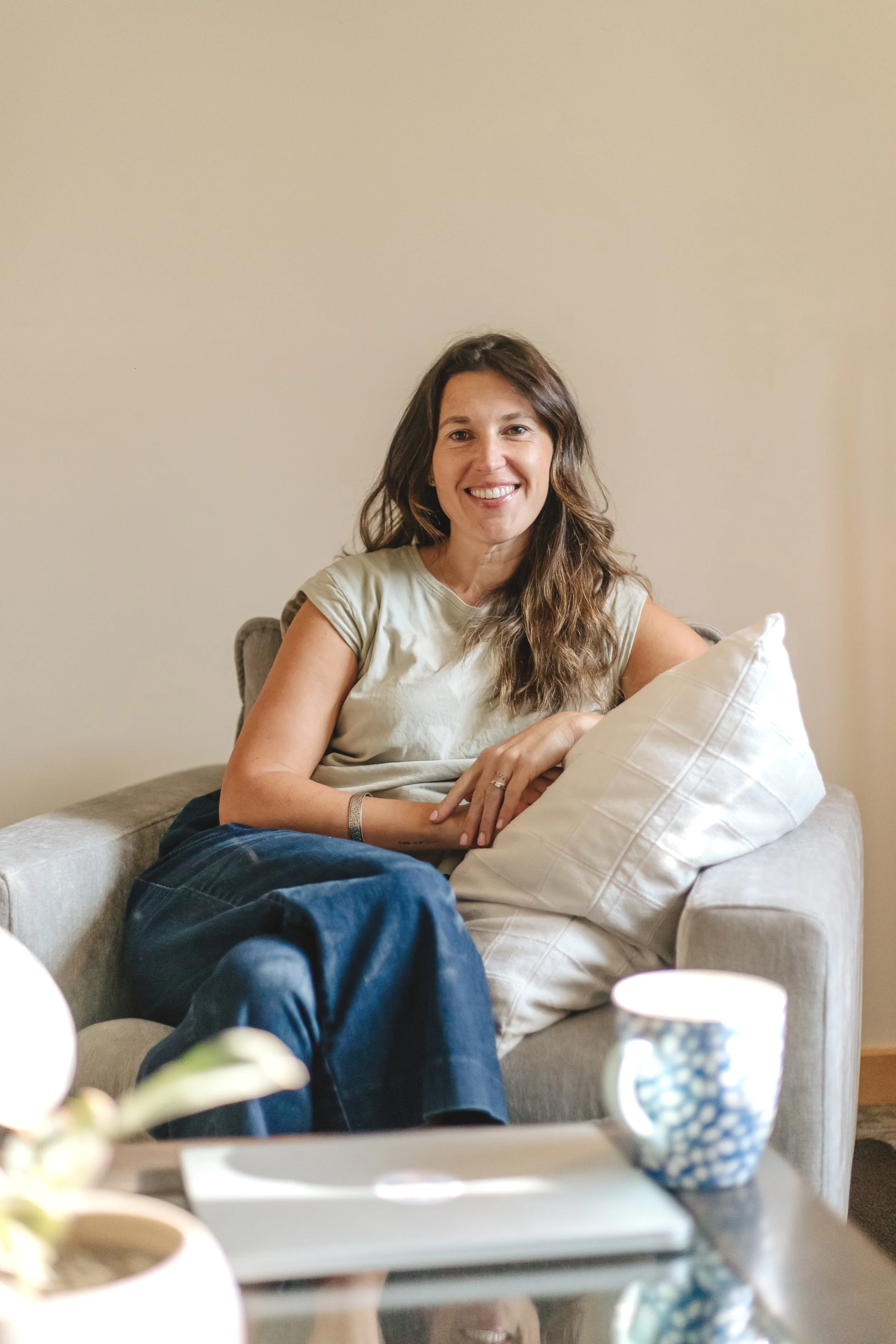 A woman with long brown hair and a beige shirt sitting on a beige couch, smiling. She has a wedding ring, a bracelet, and is resting her arms on a beige pillow. In the foreground, there is a table with a white box, a blue and white patterned mug, and a blurred flower pot.