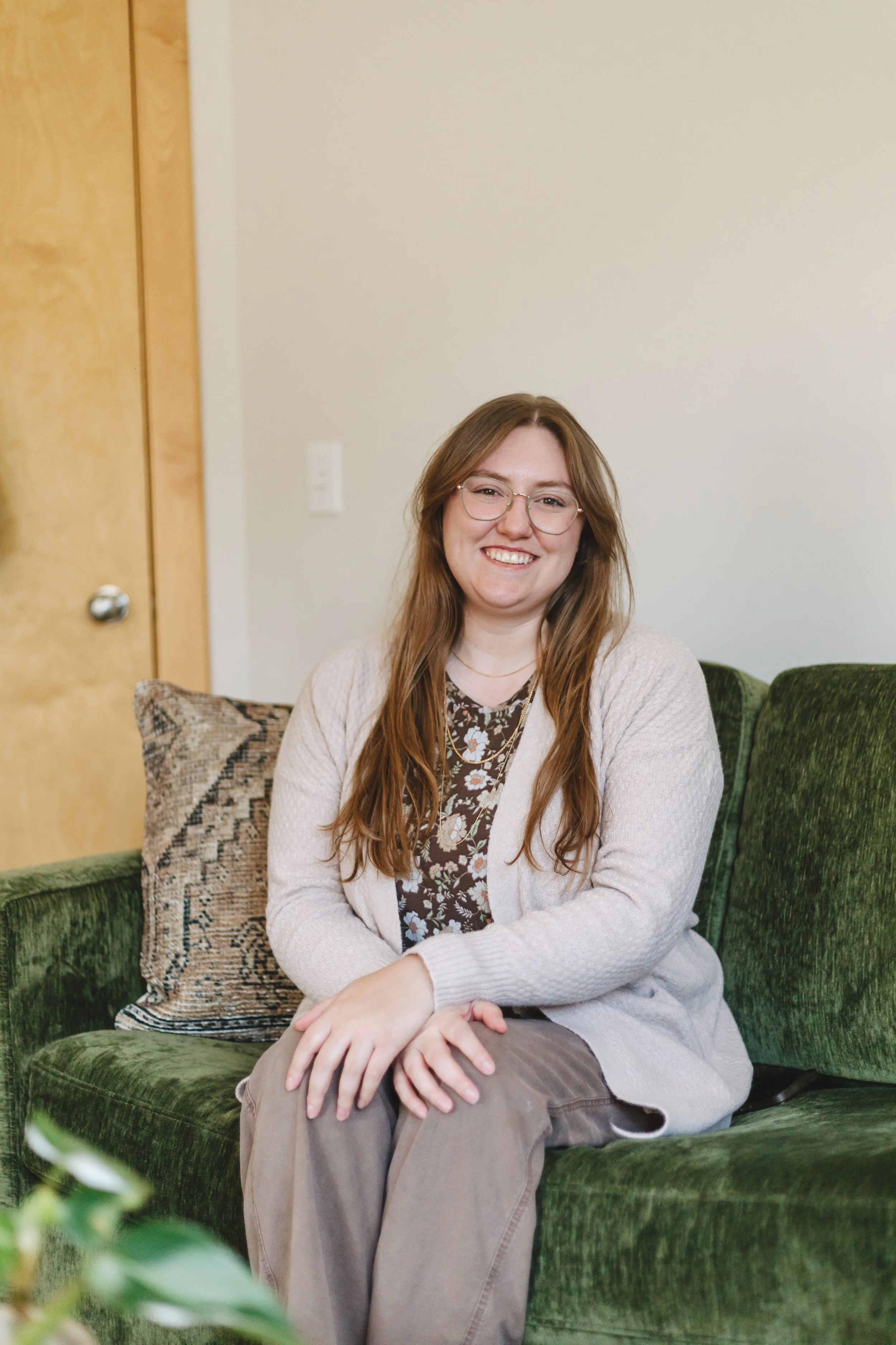 A young woman with long light brown hair and glasses sitting on a green velvet couch, smiling, wearing a patterned blouse and a beige cardigan, with pillows behind her, in a room with a wooden door, a white wall, and a leafy plant in the foreground.