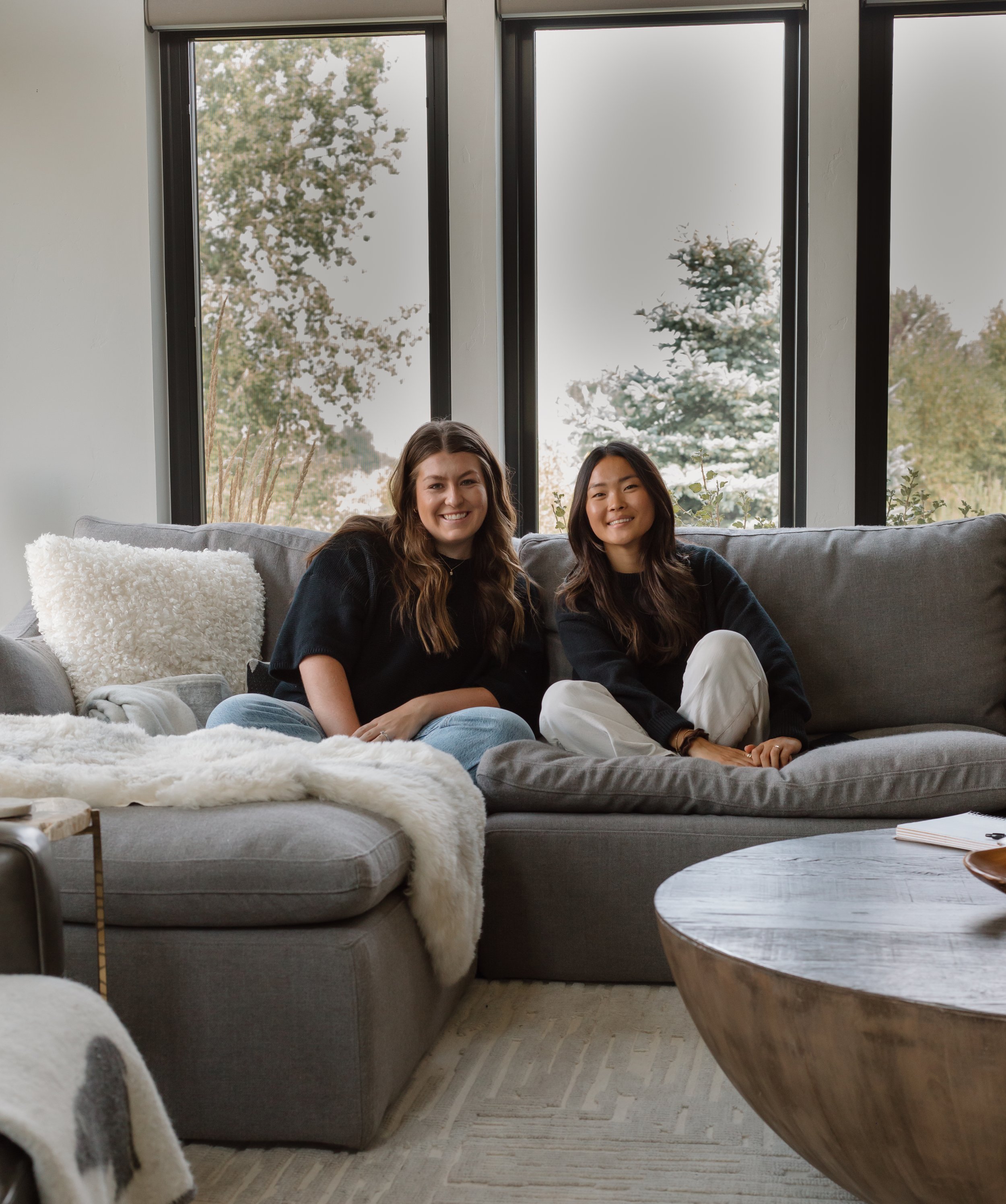 Two women sitting on a gray sofa in a living room with large windows showing trees outside, smiling at the camera.