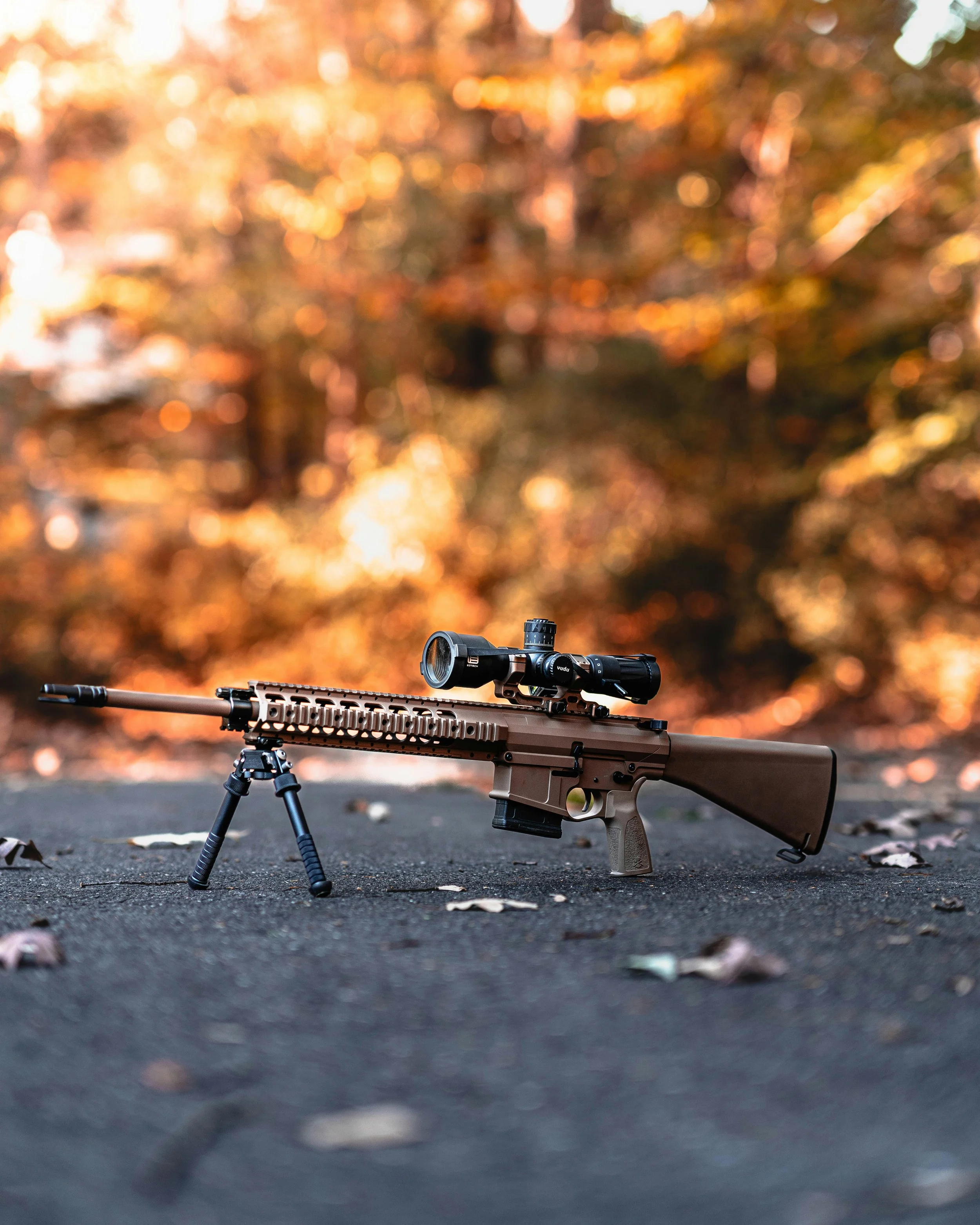 A sniper rifle with a scope and bipod placed on a rough surface outdoors, with a background of blurred autumn trees with orange and yellow leaves.
