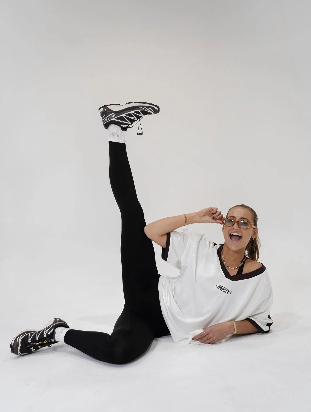 A woman wearing sunglasses, white and black sportswear, and sneakers performs a high leg stretch while lying on a white studio floor, smiling and holding her sunglasses.