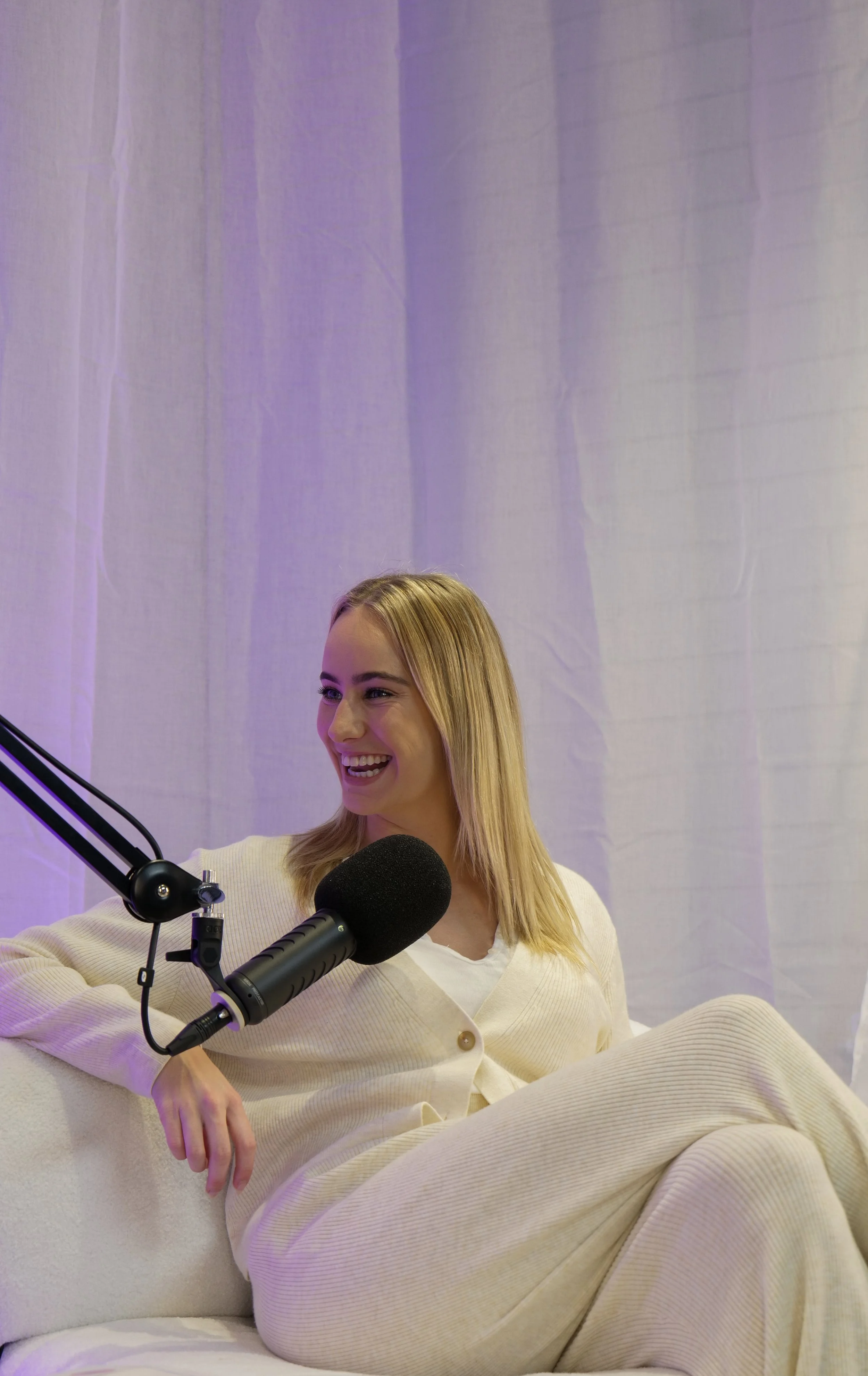 A woman with blonde hair sitting on a cream-colored sofa, smiling, wearing a cream sweater and cream pants, in front of a microphone, during a recording session. The background is a light-colored curtain with purple lighting.