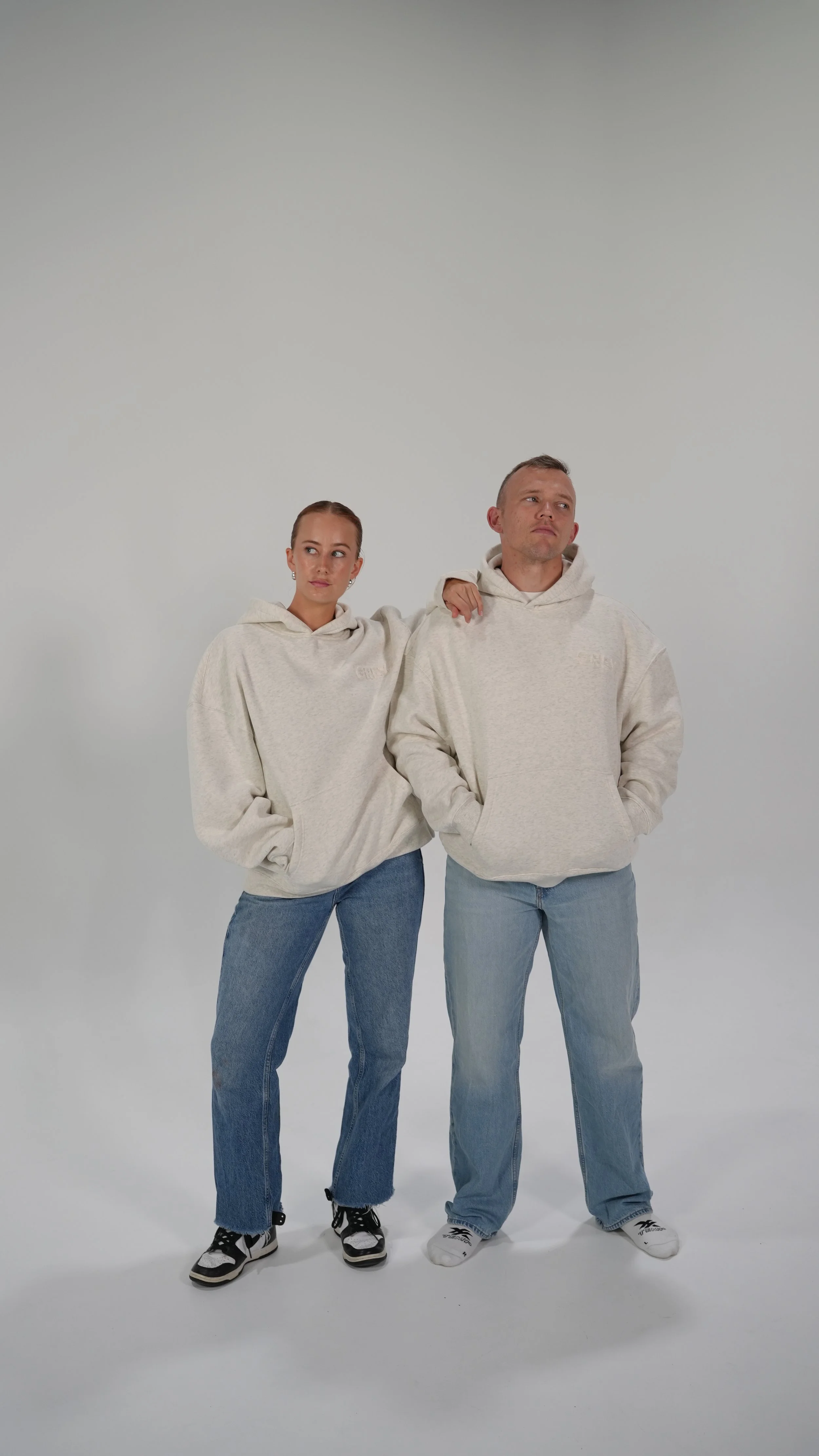 A young woman and a young man are standing side by side against a plain light gray background, both wearing cream-colored hoodies and blue jeans. The woman has her arm around the man's shoulder, and the man has his hands in his hoodie pockets. They are both looking in different directions with neutral expressions.