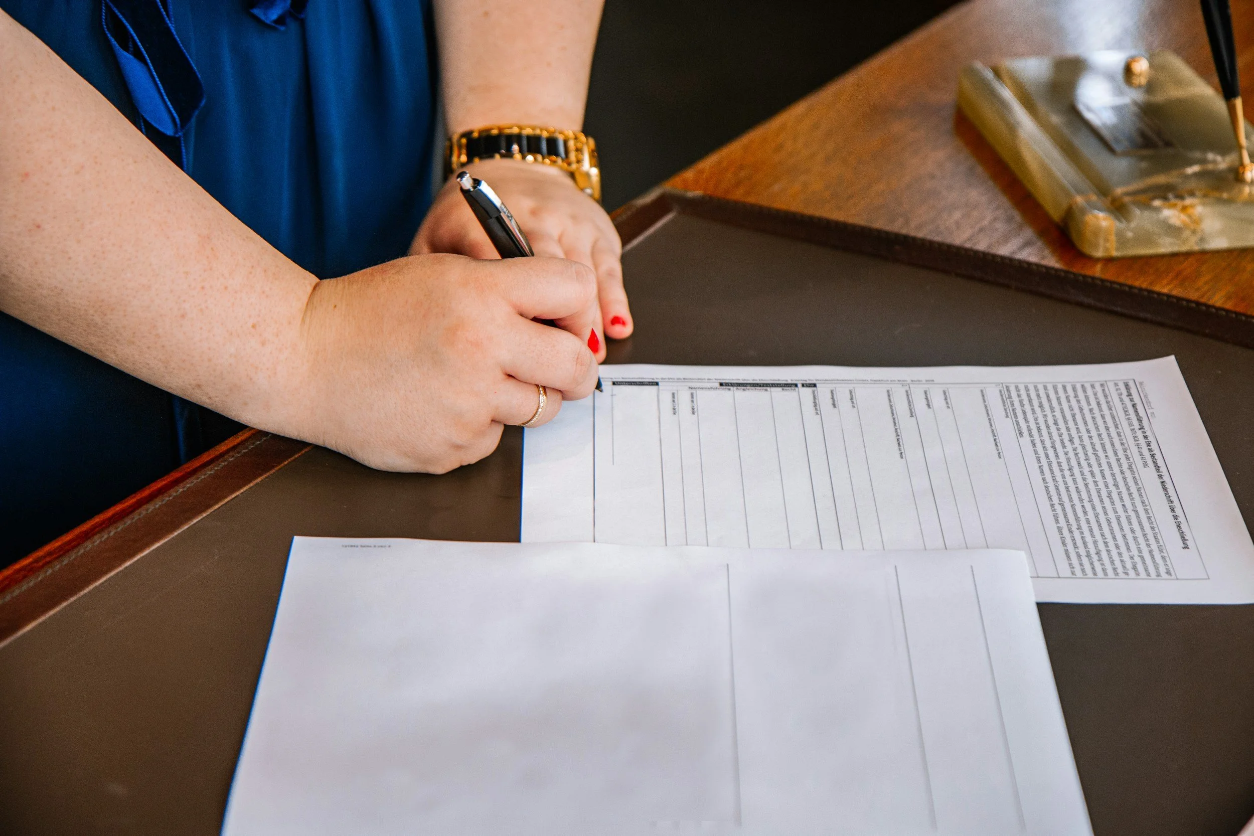 A person with red nail polish writes on a form with a black pen at a wooden desk. The person is wearing a black and gold bracelet and a blue top. A telephone and white papers are visible on the desk.
