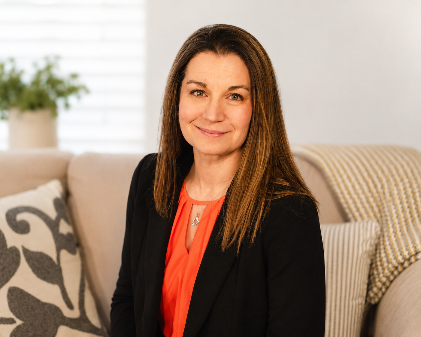 A woman with long brown hair and green eyes sitting on a beige sofa, smiling at the camera. She is wearing a black blazer and an orange blouse, with a silver necklace. The background features a window with white blinds, a potted plant, and decorative pillows.