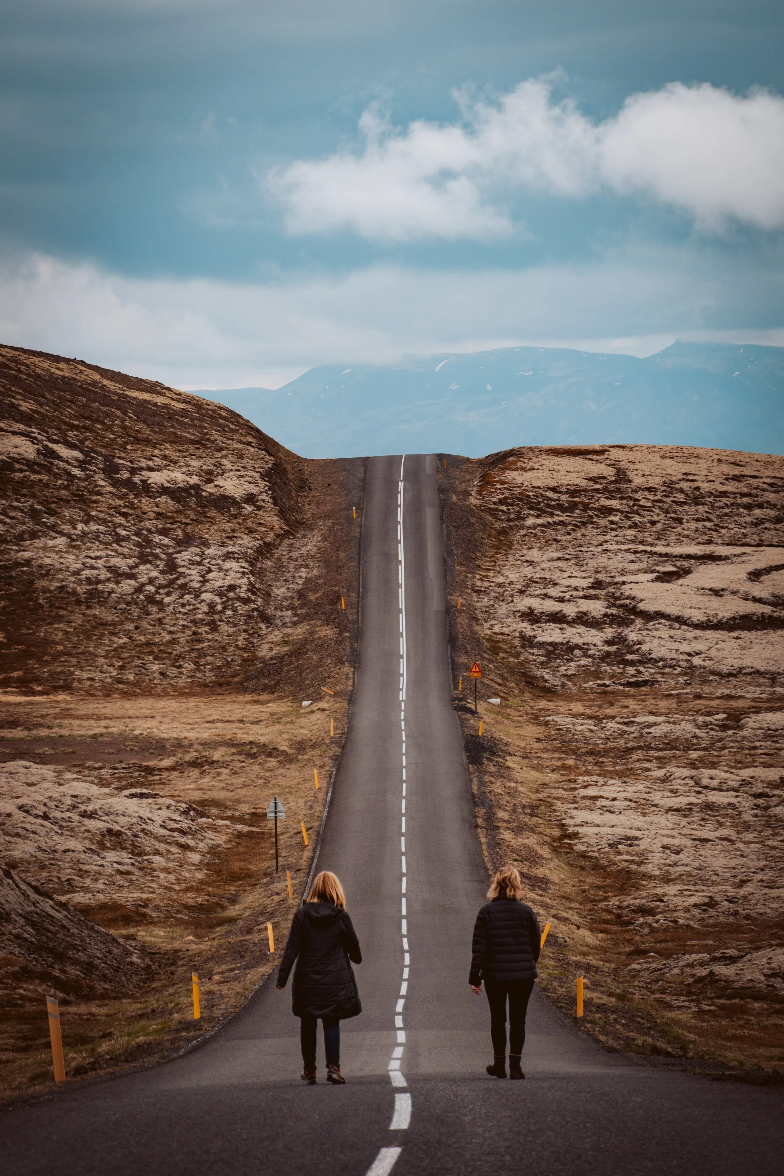 A long, straight road in a rural, hilly landscape with two people walking away from the camera, surrounded by barren, rocky terrain, under a cloudy sky.