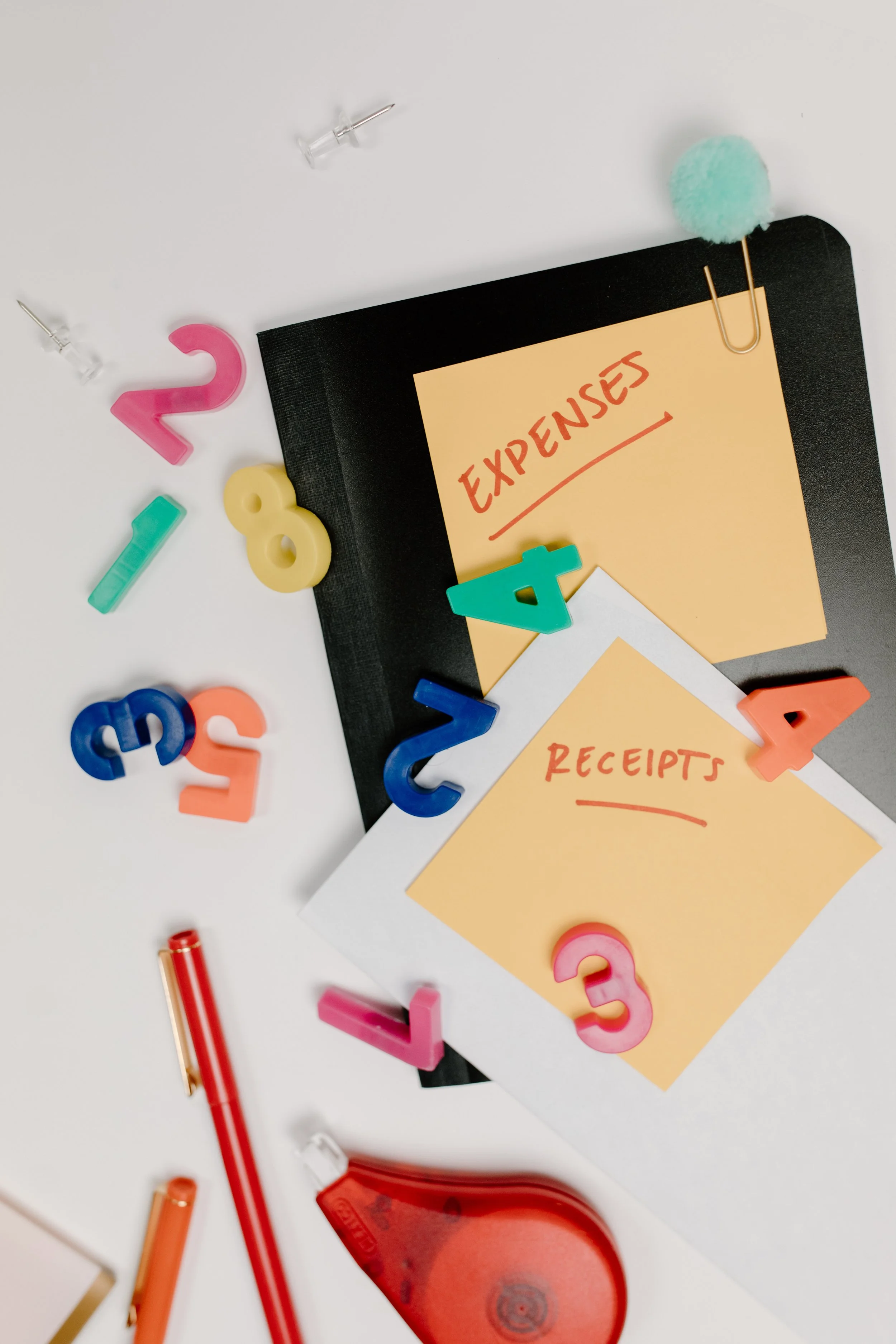 A desk with a black folder, yellow sticky notes labeled 'Expenses' and 'Receipts', colorful plastic numbers, a red adhesive tape dispenser, a red pen, a white paperclip with a pom-pom, and scattered push pins on a white surface.