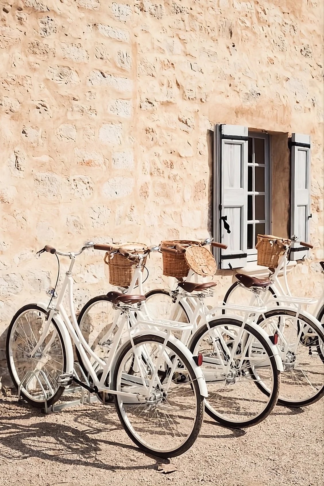 Three white bicycles with wicker baskets parked outside a stone building with a window that has white shutters.