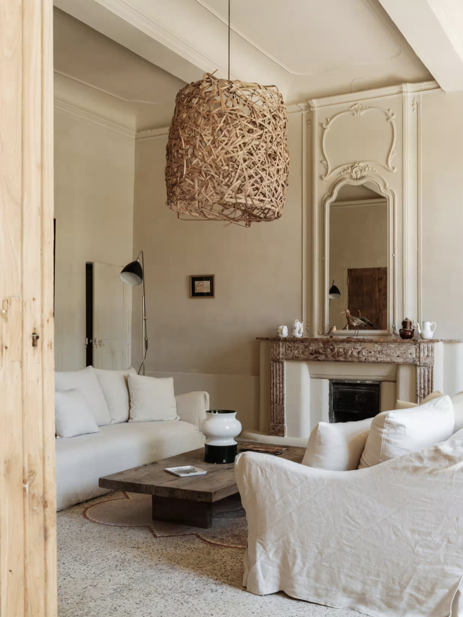 Living room with a wicker pendant light, cream sofas, a rustic wooden coffee table, a large ornate mirror above a fireplace, and decorative items on the mantle.
