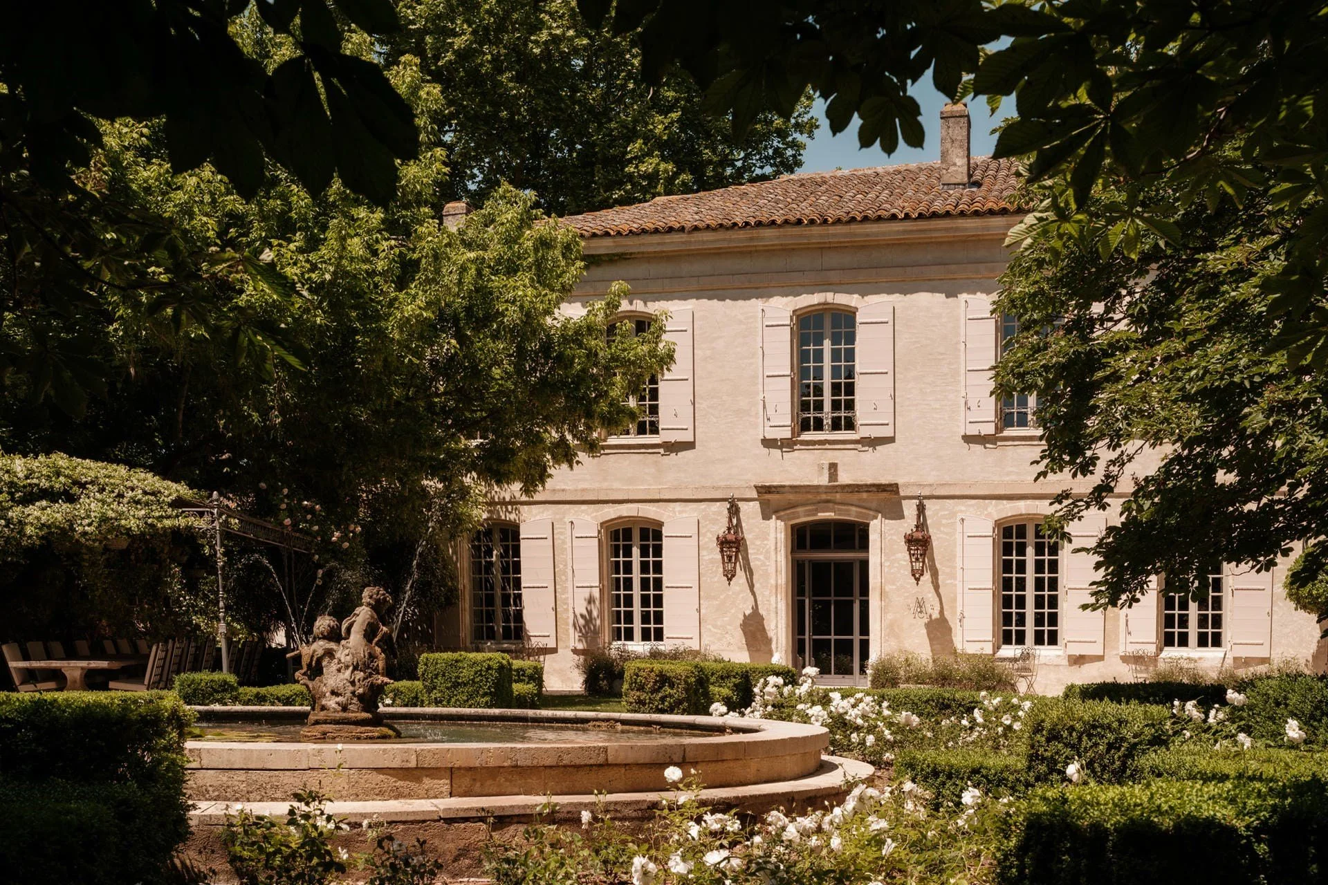 A large, elegant cream-colored house with tall windows, stone shutters, and a tiled roof, surrounded by lush trees and a well-maintained garden with a fountain and sculptures.
