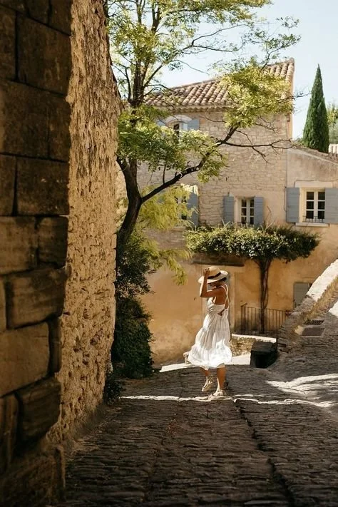 A woman walking on a cobblestone street holding a hat, with a stone house, trees, and blue shutters in the background.