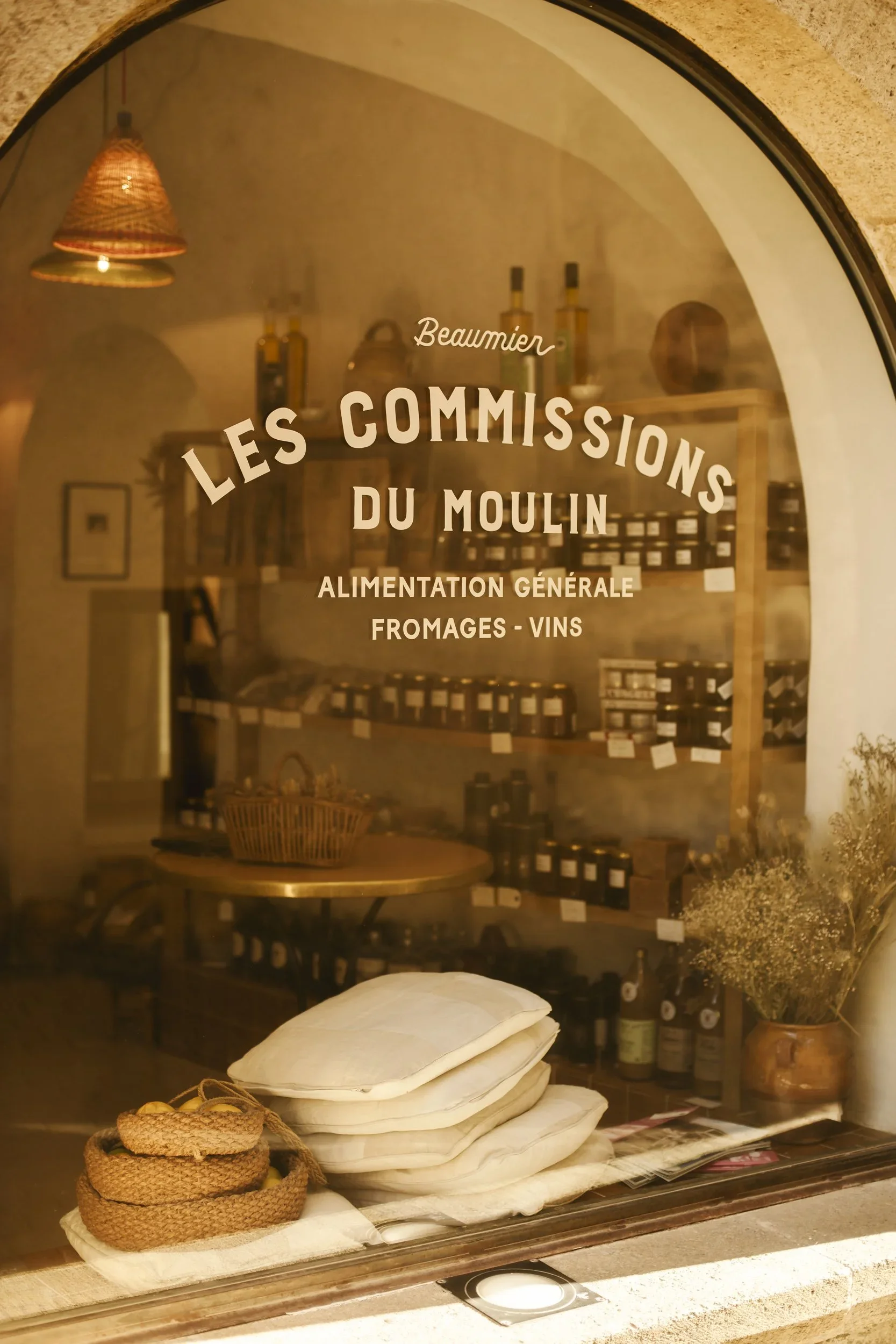 A bakery or cheese shop window with French sign saying "Les Commisisons du Moulin," indicating a focus on food, cheese, and wines. Inside, shelves hold bottles, and there are neatly stacked linens and small woven baskets on display.