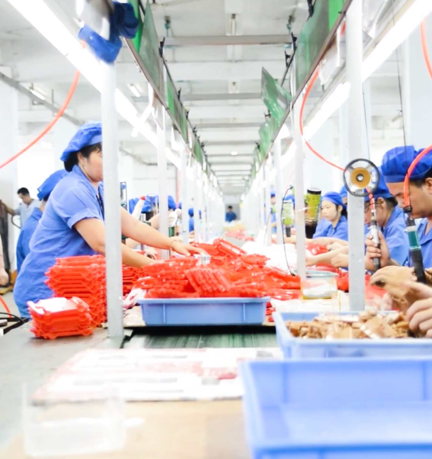 Workers in blue uniforms and hair coverings assembling red plastic components at an industrial manufacturing line.