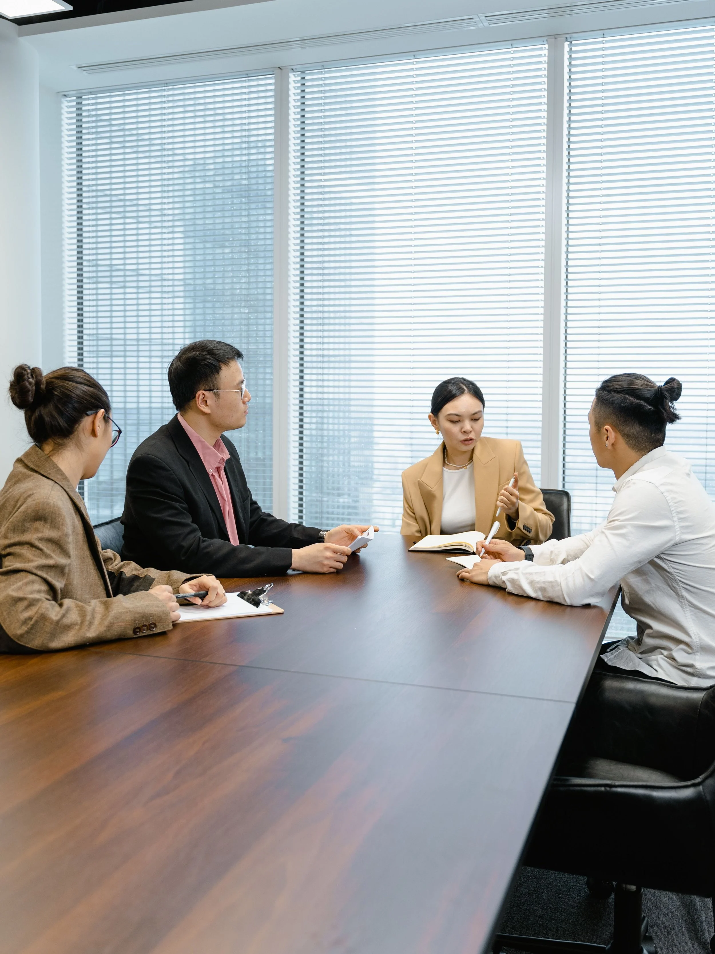 Five people in business attire engaged in a discussion around a conference table in a modern office with large windows and blinds.