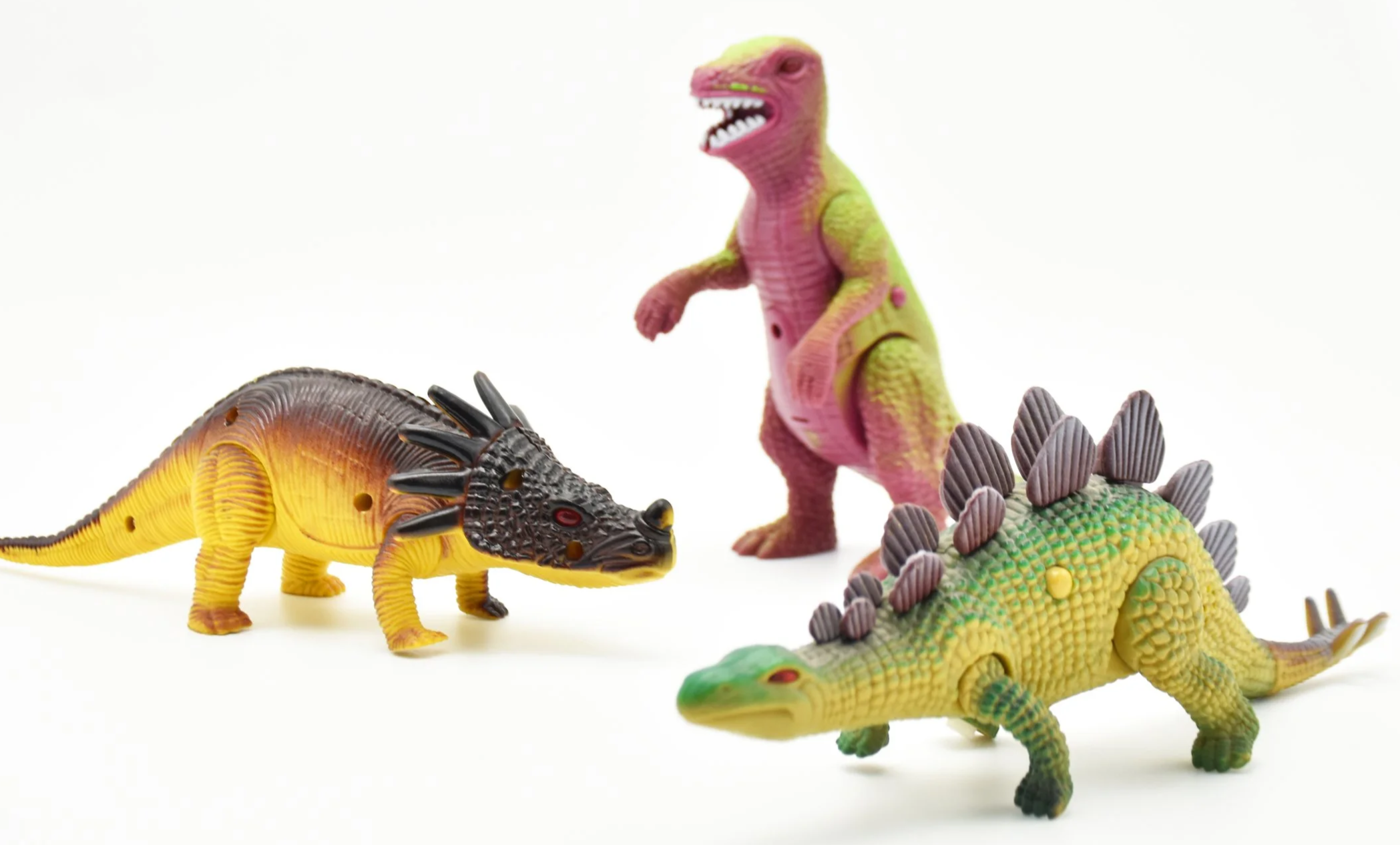 Colorful plastic toy dinosaurs including a yellow and black stegosaurus, a pink and green T-Rex, a green and yellow triceratops, and a green elongated dinosaur, all on a plain white background.
