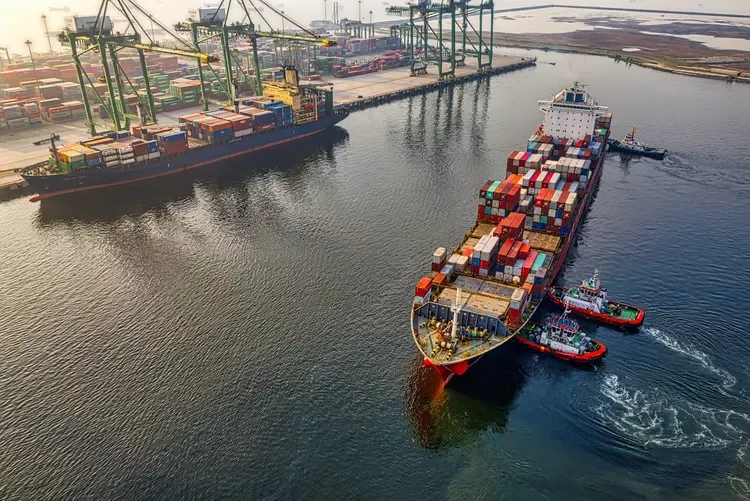 Large cargo ship with containers being guided by tugboats out of port. Container cranes and stacked containers are visible in the background.