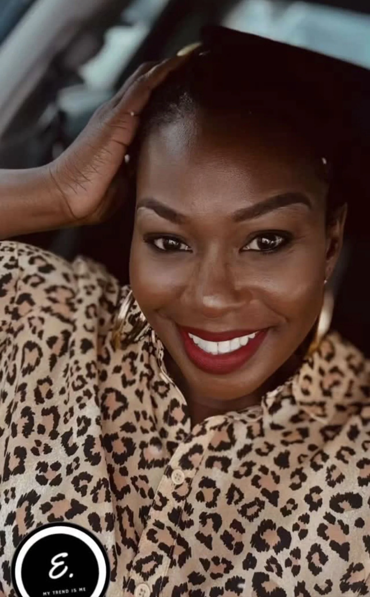 Close-up of a smiling woman with dark skin, short hair, red lipstick, and leopard print clothing, touching her short hairstyle with her right hand, indoors with a blurred background.