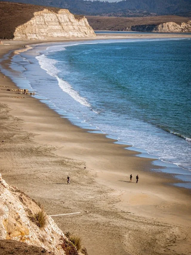 A sandy beach with a few people walking along the shoreline, facing the ocean with waves gently crashing. Cliffs and hills are visible in the background under a clear sky.