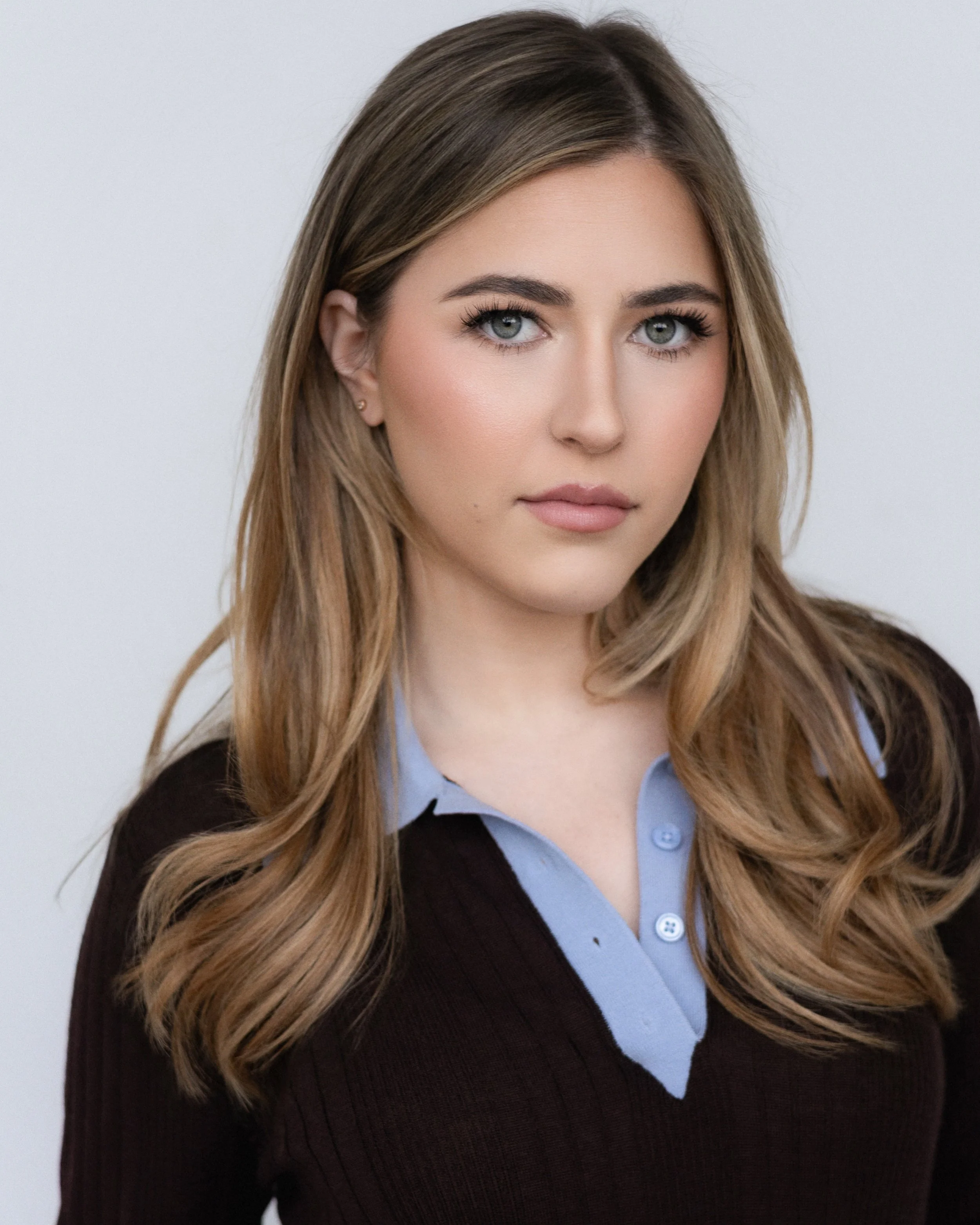 Close-up of a young woman with long, wavy light brown hair and gray eyes, wearing a light blue collared shirt layered with a dark cardigan, against a plain light background.