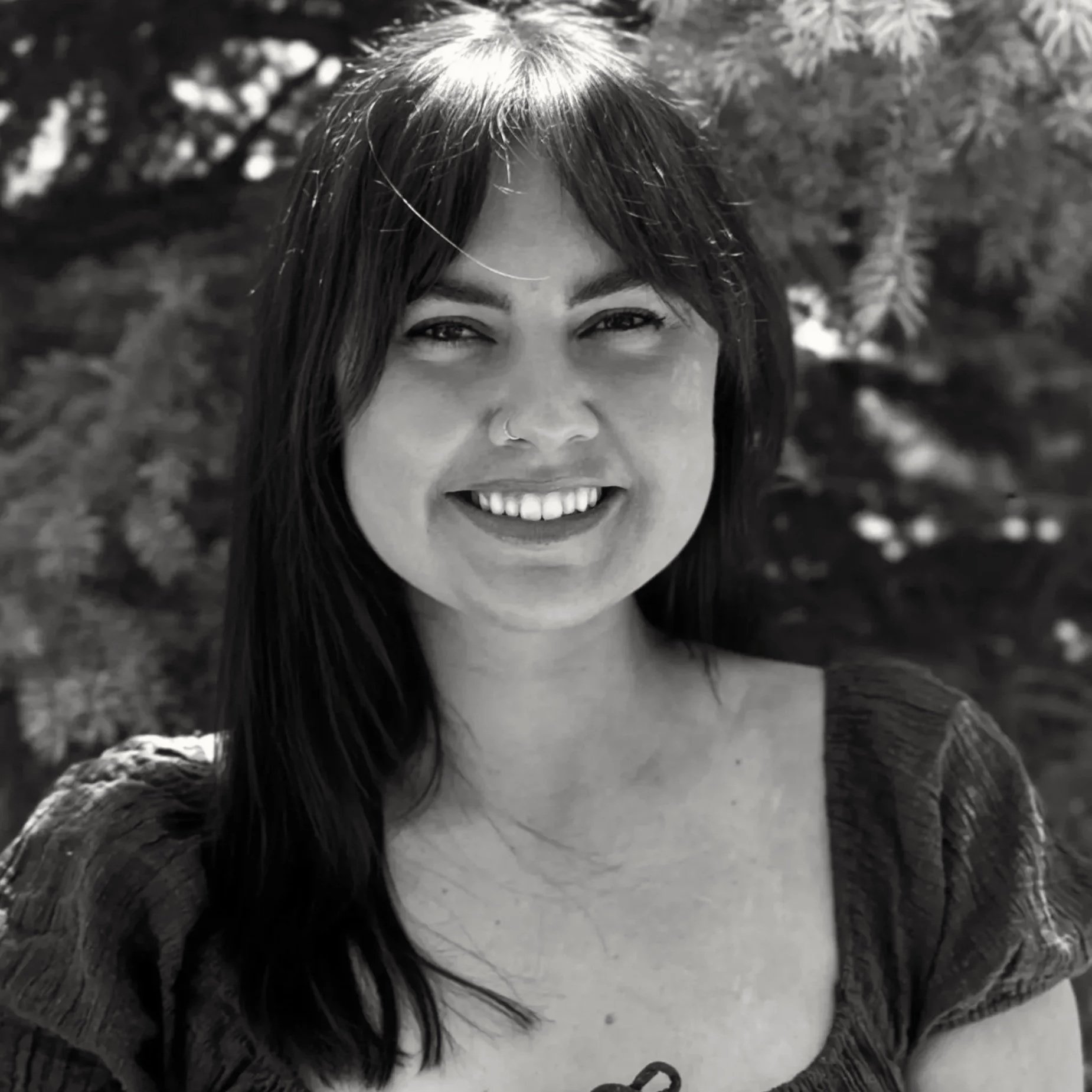 A woman with long dark hair, smiling, in an outdoor setting with trees in the background, black and white photo.