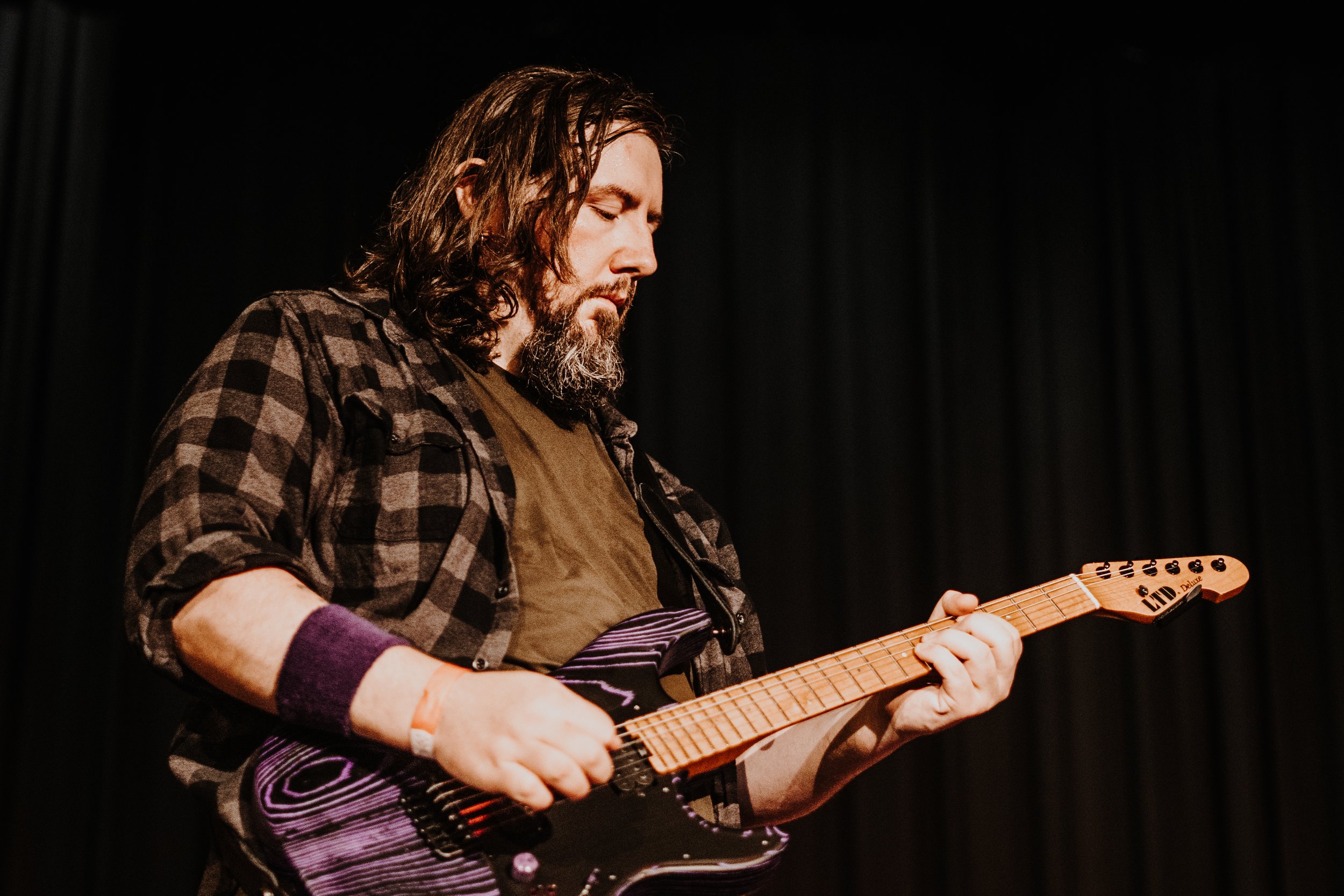 A man with long brown hair and a beard playing an electric guitar on stage, wearing a checkered shirt and a wristband, with a dark curtain in the background.