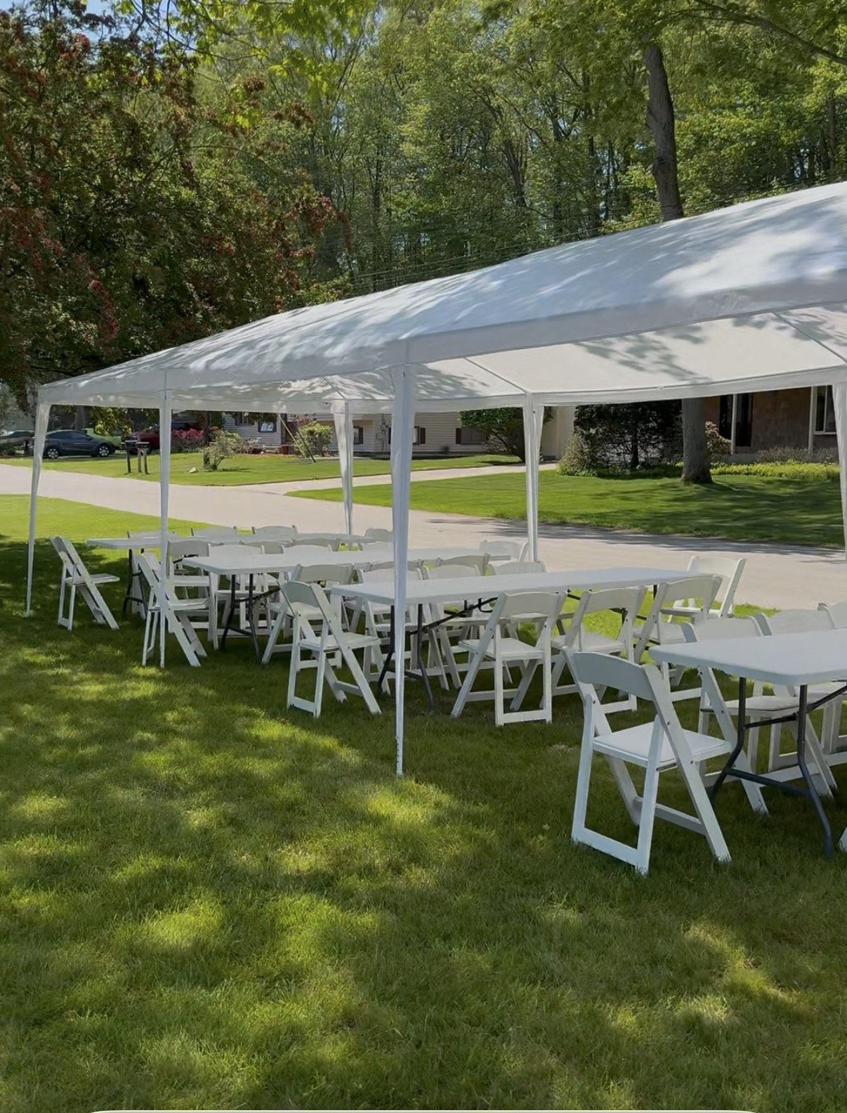 An outdoor event setup with white folding chairs and tables under a large white tent on a grassy area, surrounded by trees.