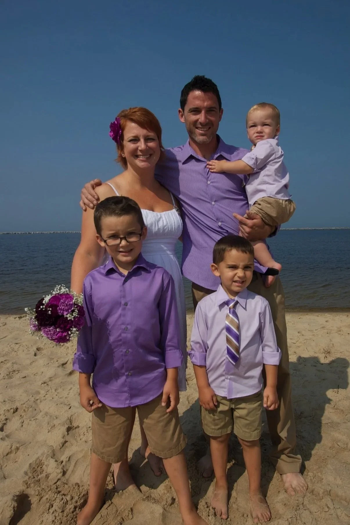 A family of six standing on a sandy beach with the ocean and blue sky in the background. The woman is holding a bouquet of purple flowers and is smiling. The man is holding a young boy who is squinting. Two boys are standing in front, both dressed in purple shirts and khaki shorts, with neutral expressions. The family is dressed in semi-formal attire, suitable for a beach wedding or special occasion.