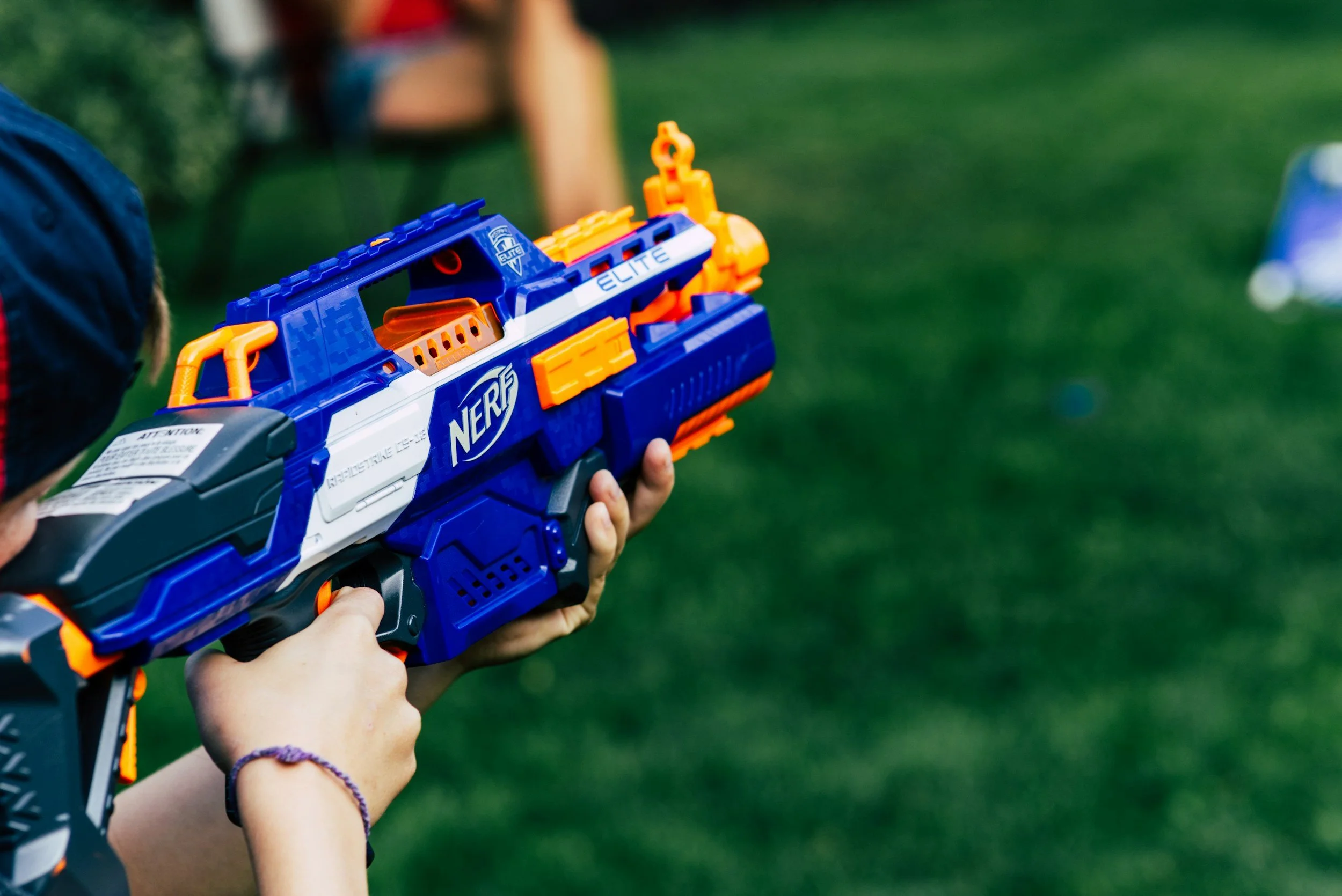 Child holding a blue, orange, and white Nerf blaster toy outdoors on grass.