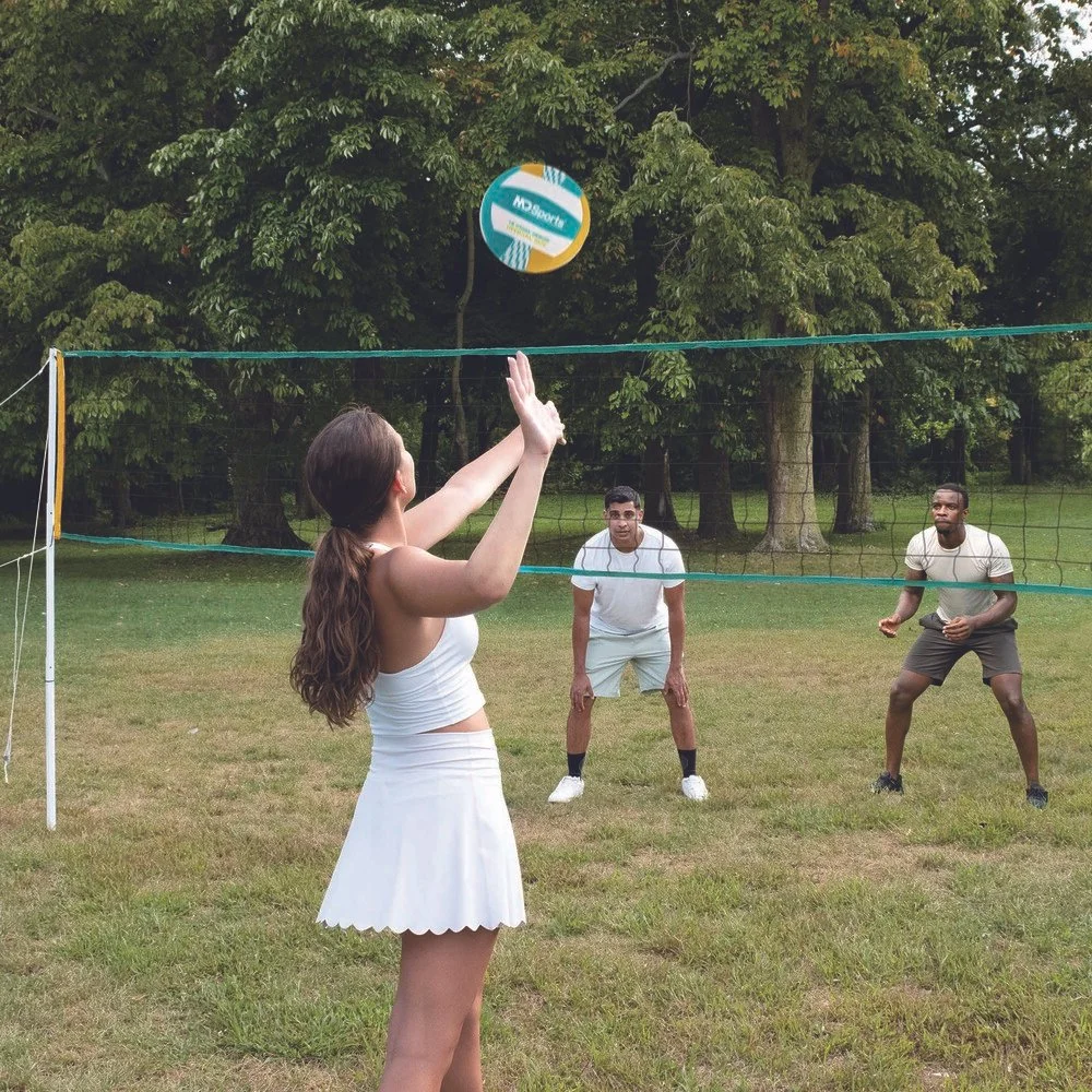 A woman playing volleyball outdoors, hitting a volleyball over the net while two men on the other side watch, surrounded by trees.