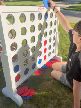 Two children playing Connect Four outdoors, with a large white Connect Four game on the grass.