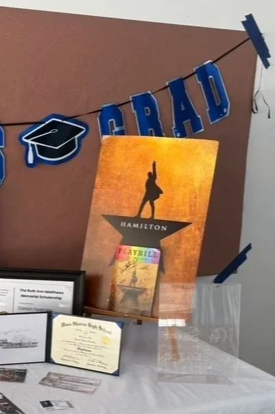 A graduation decoration with a banner spelling 'GRAD' and a graduation cap graphic, a Hamilton musical poster, and a playbill on a table.
