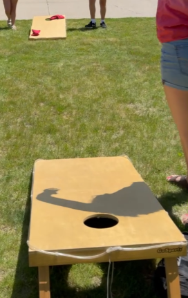 A person wearing a pink shirt and shorts is about to toss a bean bag on a yellow cornhole board set up on grass, with another cornhole board and player visible in the background.
