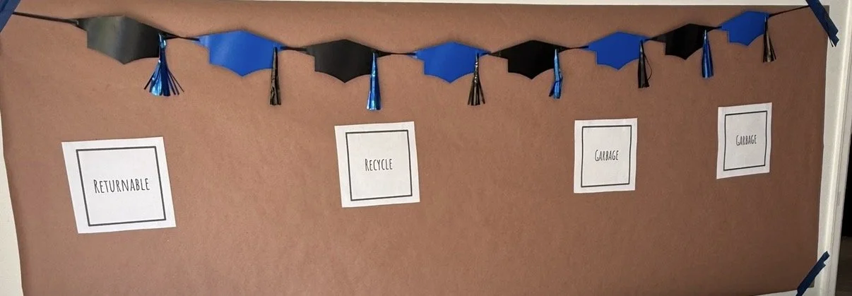 Bulletin board decorated with a blue and black tassel garland, with four labels reading 'Returnable,' 'Recycle,' 'Garage,' and 'Garbage' attached to the board.