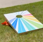 Colorful rainbow-themed cornhole game on grass with a small orange cone nearby.