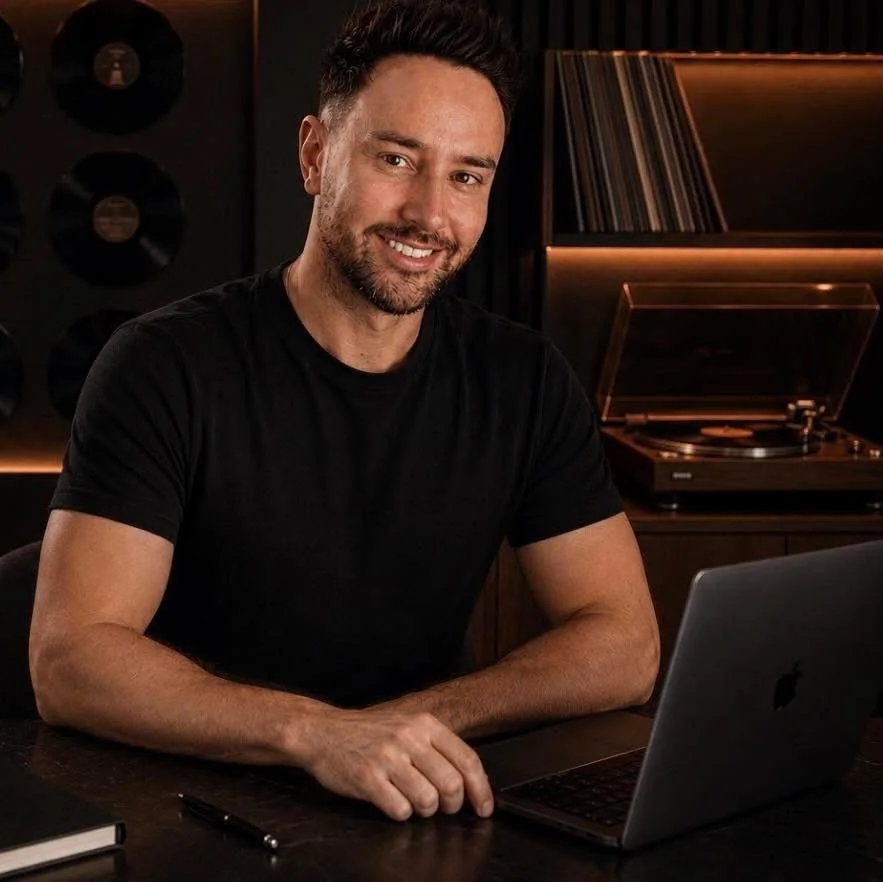 A man with dark hair and a beard sitting at a desk with a laptop, smiling at the camera. In the background, there are vinyl records and a vintage turntable.