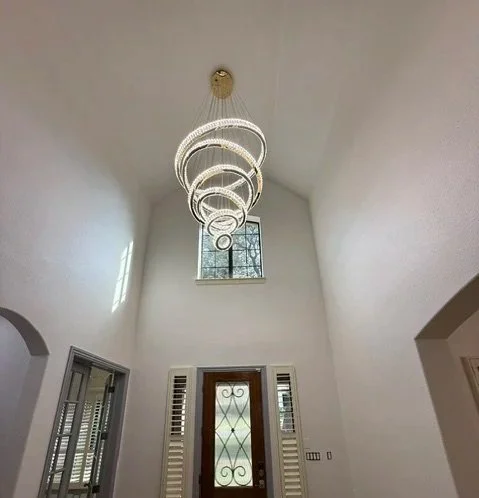 Modern chandelier hanging from a high ceiling in an entryway with a decorative front door and tall window.