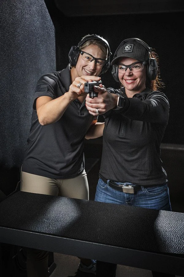 Two women wearing ear protection and glasses at a shooting range, practicing aiming a handgun together.