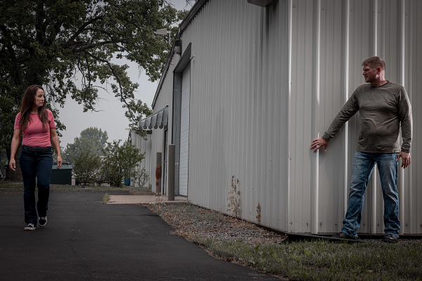 A young woman in a pink shirt and jeans walking on a paved path, looking at a man in a gray shirt and jeans standing against a large metal building, touching the wall with his hand.