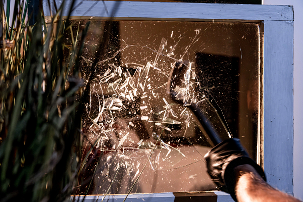 A person wearing black gloves breaking a glass door with a hammer, causing shattered glass pieces to fly outward.
