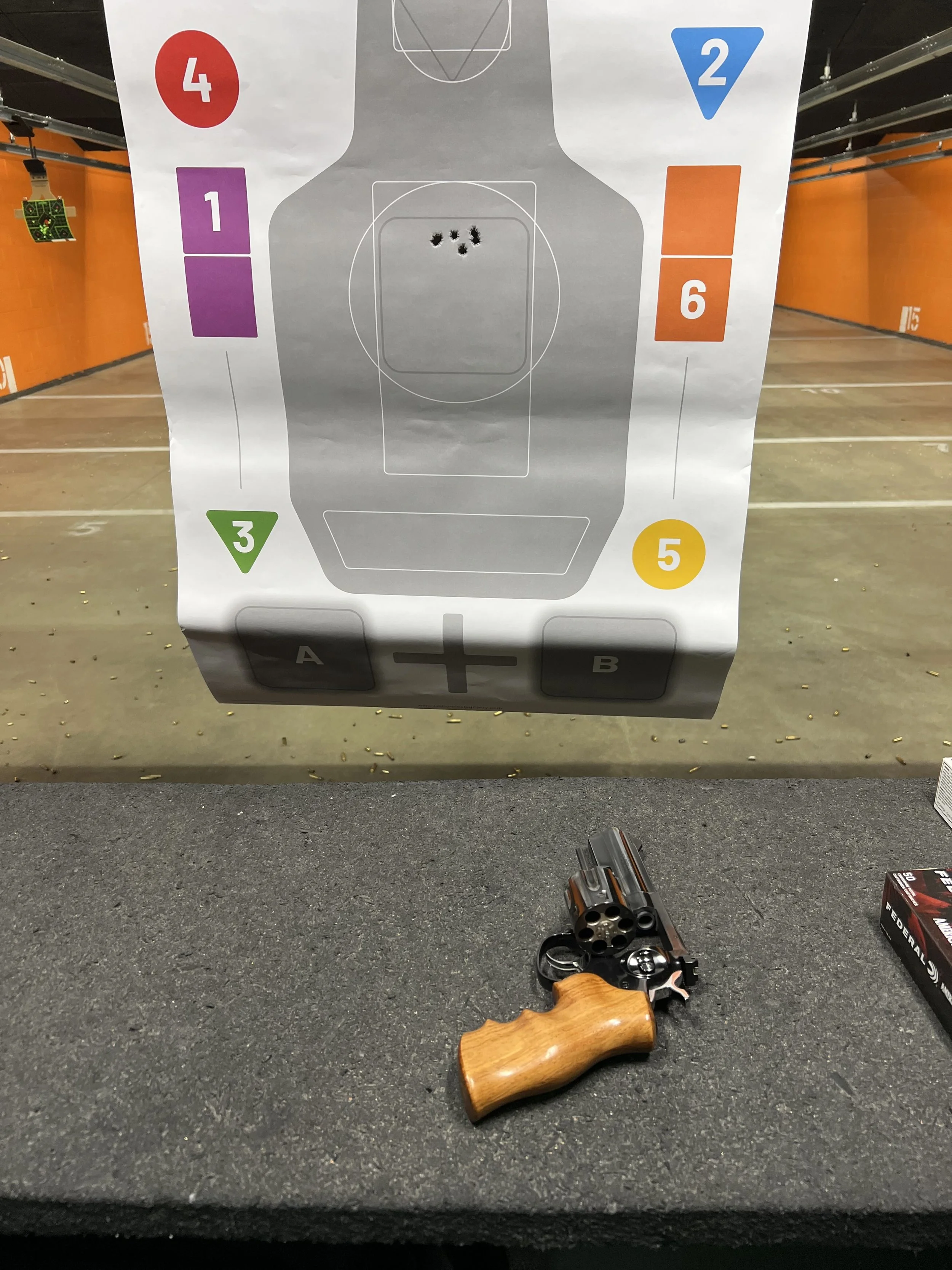 A target shooting paper with colored zones numbered 1 to 6, a revolver with a wooden grip and four bullets on a black surface, and a box of ammunition in the background.