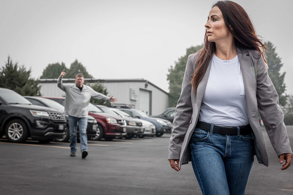 Woman walking in a parking lot with cars, looking away, while a man in the background waves his arms.