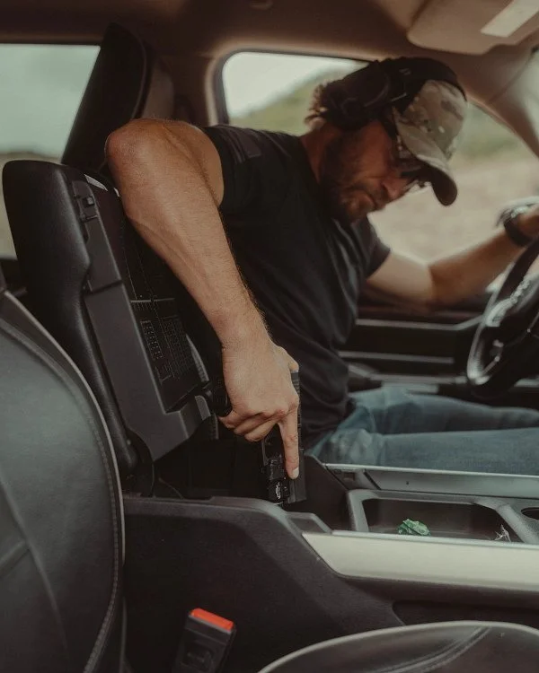 A man in a black t-shirt and camouflage cap sitting in a vehicle, inserting a firearm into a compartment between the seats.