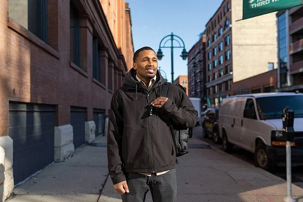 A man in a black jacket walking on a city sidewalk with buildings, parked cars, and a streetlamp in the background.