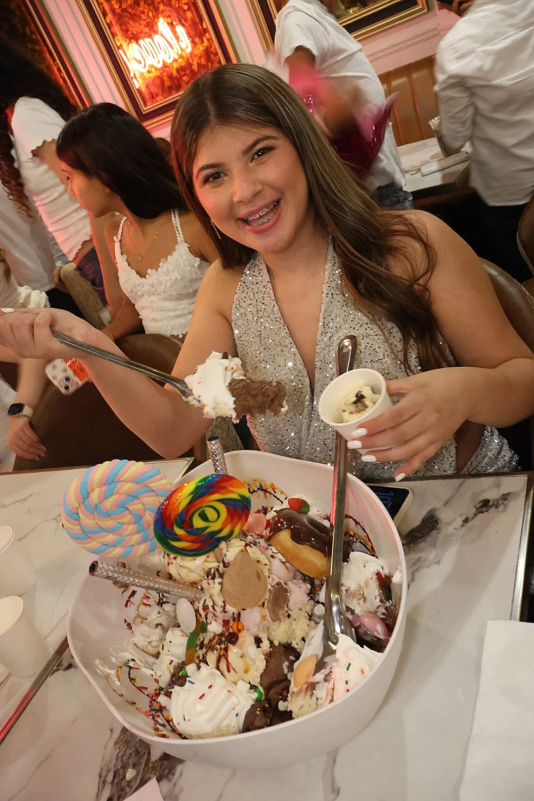 Young woman at a celebration, smiling and holding a spoon near a large bowl of ice cream with colorful candies, a lollipop, and whipped cream. Other people are in the background in a decorated, elegant setting.