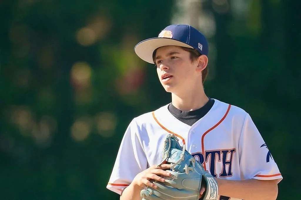 Young boy playing baseball, wearing a white jersey with orange and navy accents, a navy cap, holds a baseball glove, outdoor setting with trees in the background.